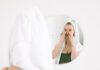 A woman in a towel performing her skincare routine in front of a bathroom mirror.
