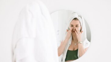 A woman in a towel performing her skincare routine in front of a bathroom mirror.